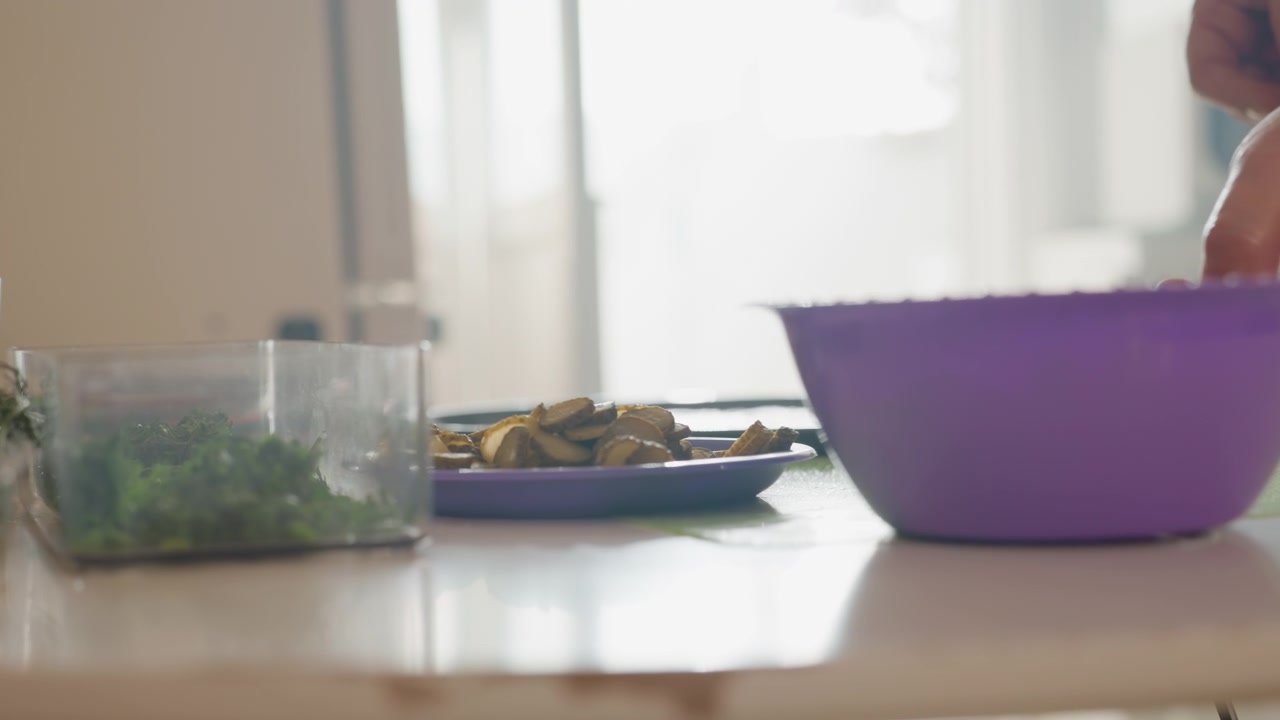 Low angle view shows hand reaching toward small plate beside purple bowl on light wooden table, with soft daylight, natural shadows, and gentle motion in cozy home dining setting, with hand, reaching