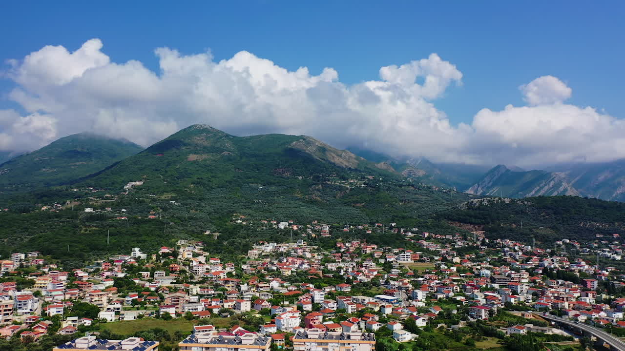 Drone footage above the multiple houses built at the foot of beautiful mountains in Bar, Montenegro. Huge white cloudscape at backdrop in the blue sky.