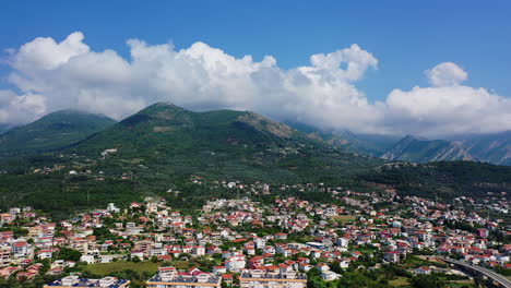 drone footage above the multiple houses built at the foot of beautiful mountains in bar, montenegro. huge white cloudscape at backdrop in the blue sky.