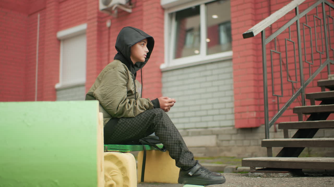 Side view of student in hooded jacket seated outdoors playing game on handheld device with focused expression, urban background of red brick building, stair railing