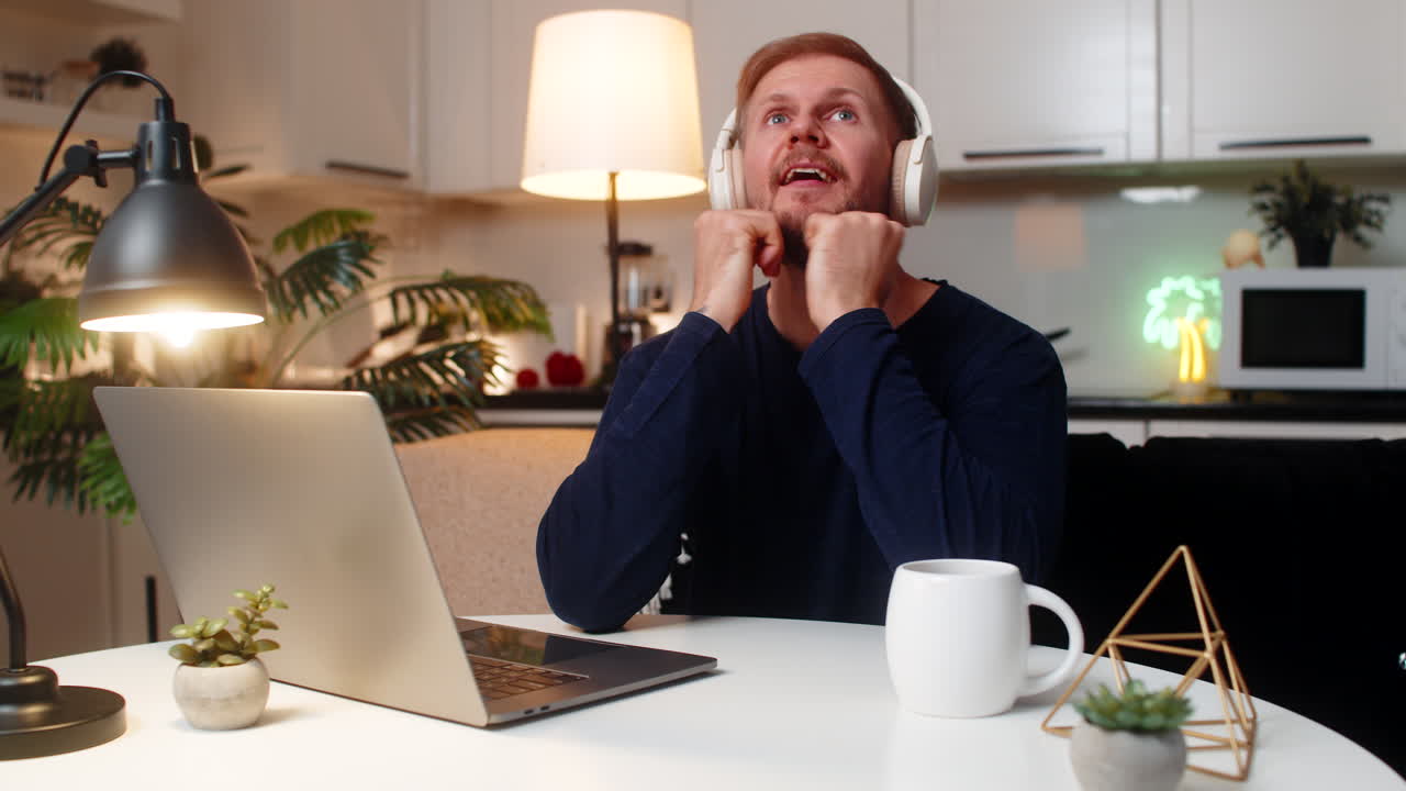 Giovane uomo con cuffie wireless sogna e pensa mentre lavora al portatile concentrato nell'atmosfera serale