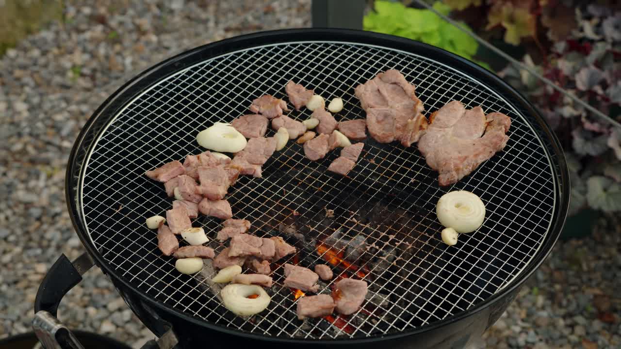 Using tongs, a woman places a fresh slice of raw marbled beef steak from a tray onto a hot charcoal grill to cook with pork and garlic at an outdoor garden barbecue