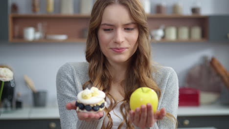 Portrait-of-attractive-woman-choosing-between-cake-and-apple-at-home-kitchen.