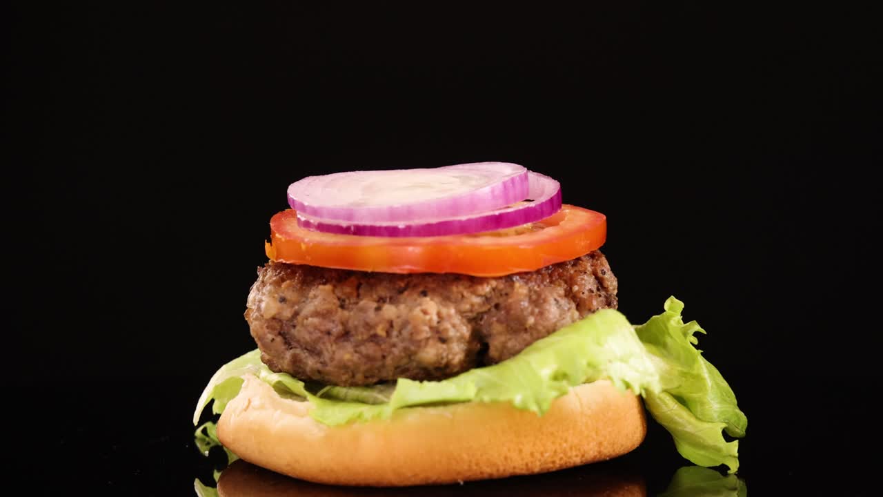 A hand places a red onion slice onto a tomato-topped beef burger with lettuce on a bun, shot against a black background with bright, even lighting