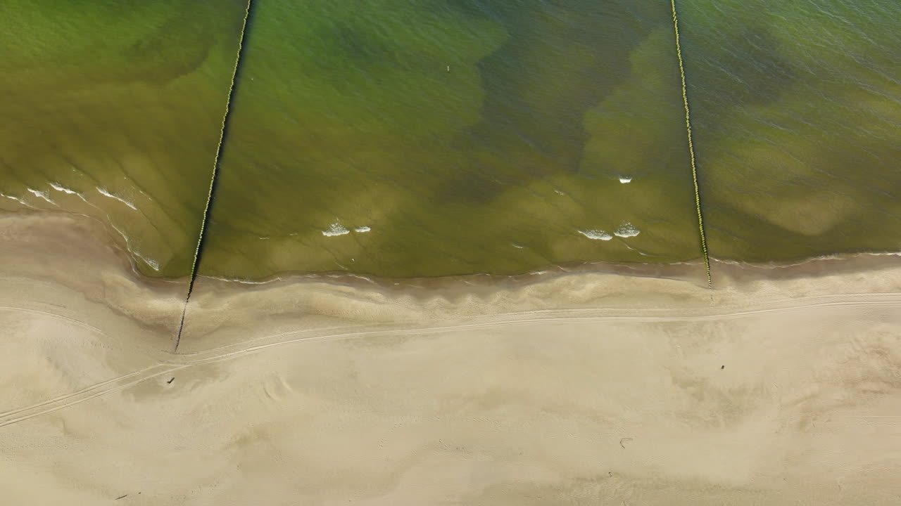 Top-down aerial view capturing the intersection of a sandy beach and shallow green sea waters, segmented by narrow groynes extending into the sea - horizontal