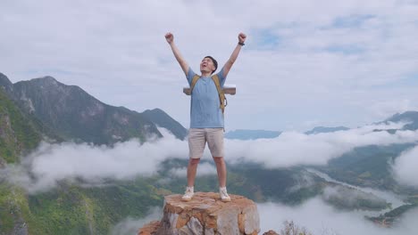 asian hiker male standing on the rock and raising his hands celebrating reaching up top of foggy mountain