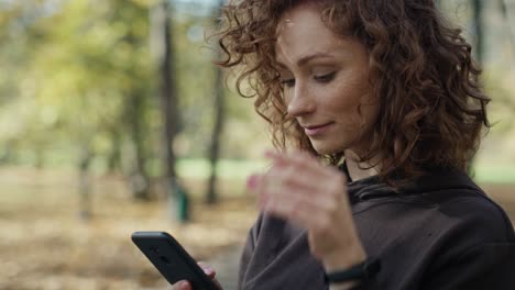 Mujer-Jengibre-Sonriente-Parada-En-El-Parque-Y-Usando-El-Teléfono-Móvil