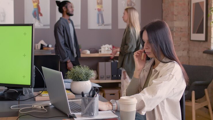 Mujer concentrada sentada en el escritorio y trabajando en la computadora en la oficina mientras dos colegas multiétnicos hablan entre sí