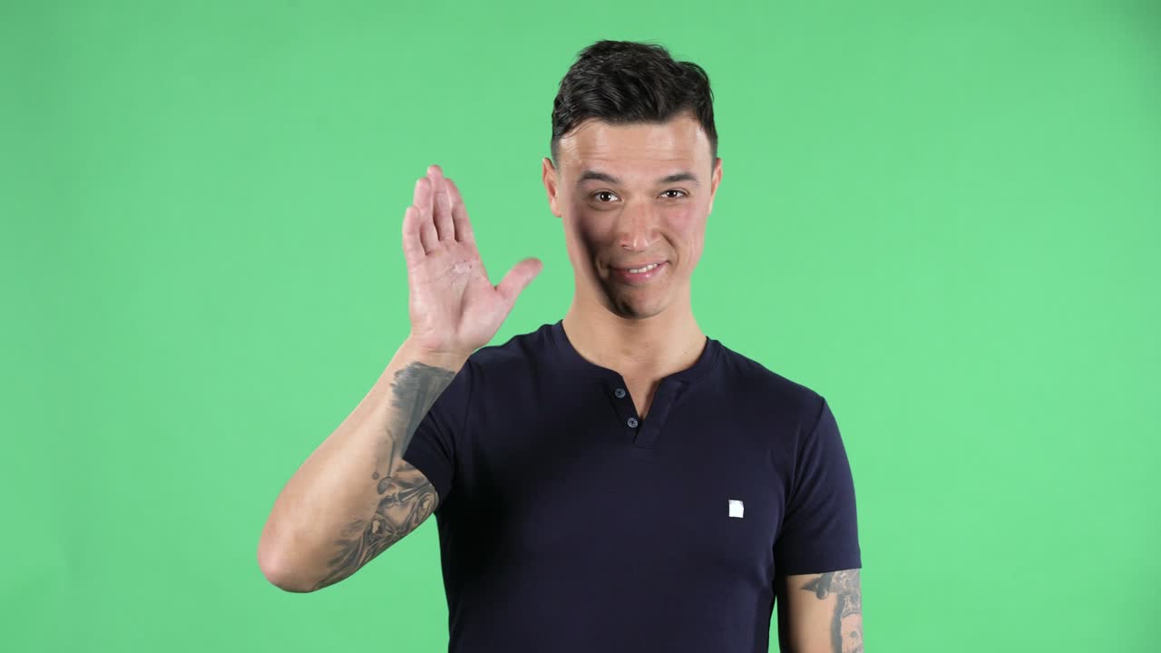 Portrait of handsome young guy is waving hand and showing gesture come here. Burning brunette with stylish hair in a black T shirt and tattoos on his hands on a green screen in a studio