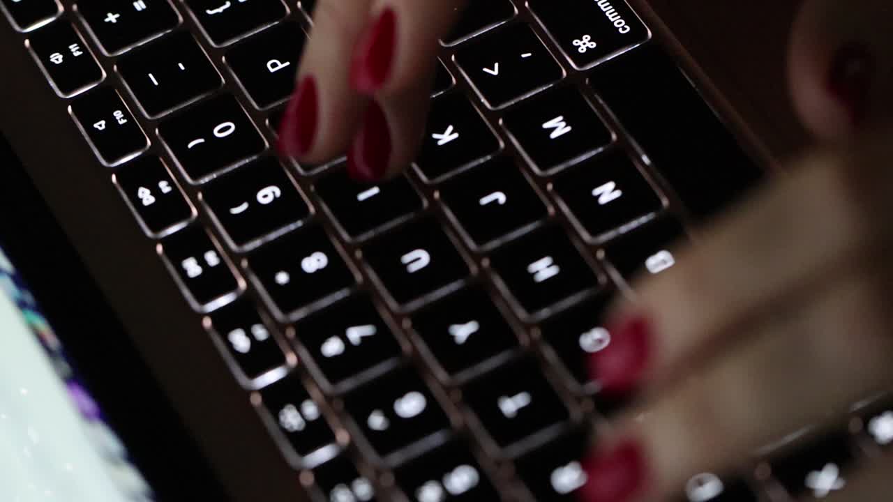 Close-up of hands with red nail polish typing on a backlit keyboard