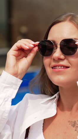 happy relaxed young lady in white shirt sitting near the pool. woman puts on her sunglasses and smiles to the camera. vertical video
