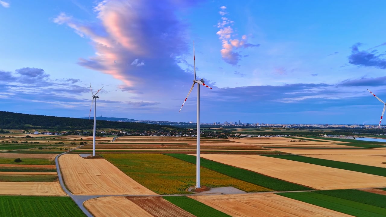 Vindmøller på farverige marker nær en fjern by. Droneudsigt over alternative energigeneratorer over grøn og gylden jord under en overskyet solnedgangshimmel.