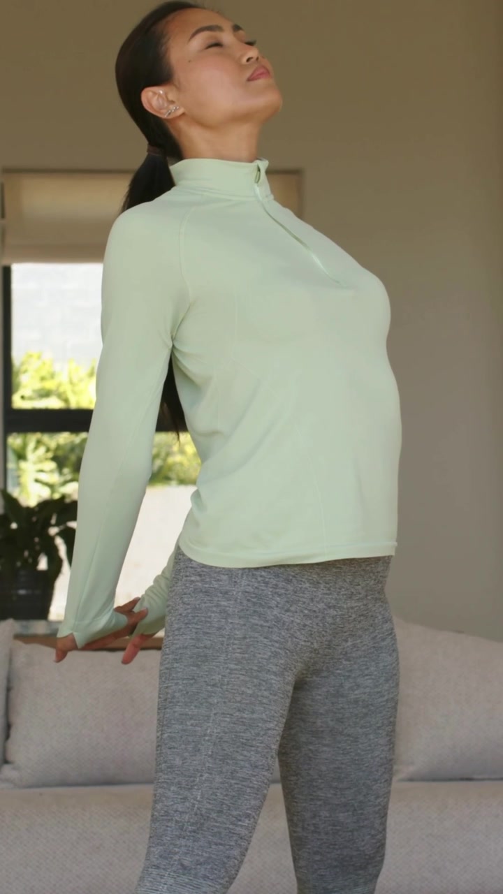 Vertical video: Starting woman in green zip-up using sofa at home, extending arms, stretching