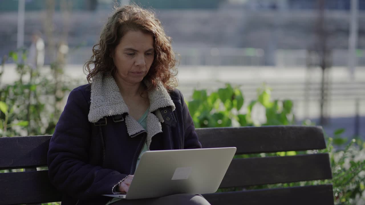 Thoughtful mature woman typing on keyboard of laptop outdoor