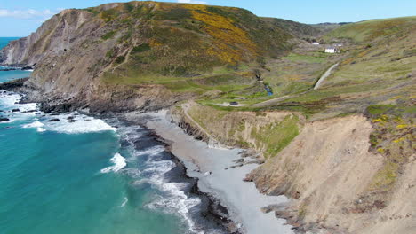 Aerial-coastal-shot-of-the-waves-in-the-sea-at-Spekes-Mill-beach-in-Devon-near-a-carpark-and-green-hills