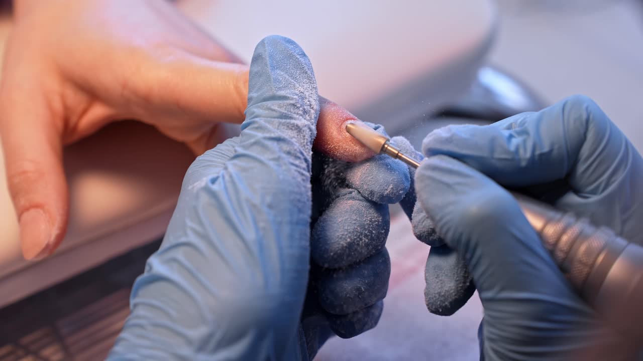 Primer plano de salón de una técnica de uñas refinando una uña natural con una lima eléctrica, capturando el polvo, la preparación precisa de la manicura y el flujo de trabajo profesional del cuidado de uñas.