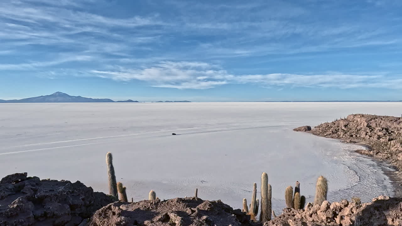 Panorâmica das salinas da Bolívia com um carro passando sob montanha coberta de cactos