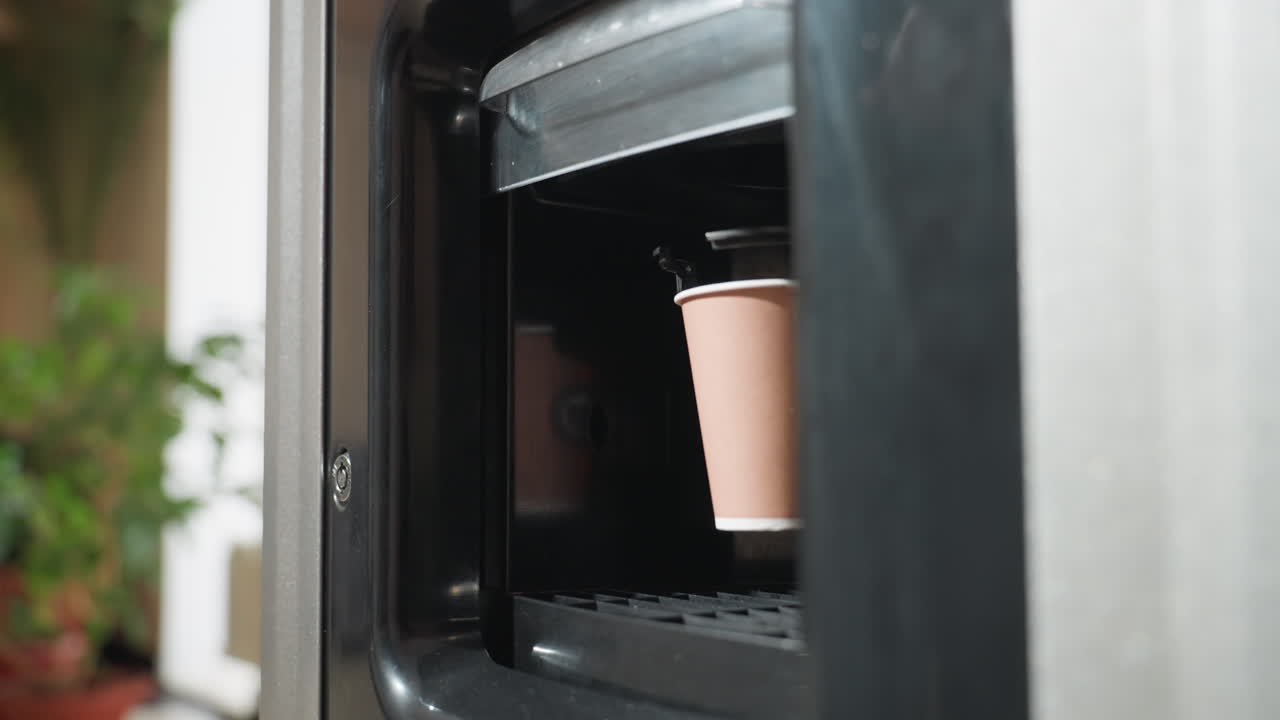 자판기에서 뜨거운 커피가 담긴 종이컵이 나오는 모습이에요. 컵 배출구가 잘 보이고, 현대적인 사무실 배경에 흐릿하게 보이는 식물이 있어 따뜻한 음료가 제공되는 분위기를 연출해요.