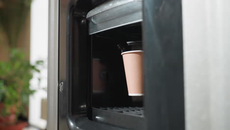 paper cup receiving hot coffee inside vending machine dispenser with visible cup drop area, capturing warm beverage being served in modern office setting with blurred plant in background