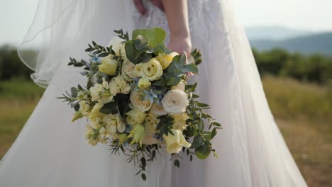 novia de pie sosteniendo su ramo de flores de boda en el campo de hierba afuera después de la ceremonia en vestido blanco tiro de cardán de primer plano en el día nublado de verano