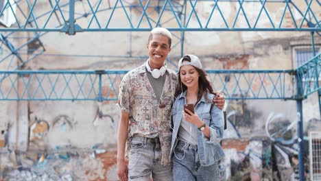 Smiling-man-and-his-caucasian-girlfriend-wearing-headphones-and-watching-something-on-the-smartphone-while-walking-down-the-street