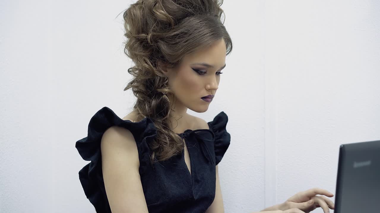 사무실에서 노트북에 앉아 텍스트를 타이핑하는 젊은 소녀는 때때로 전화를보고