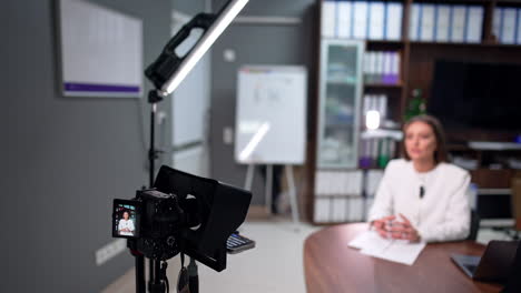 caucasian brunette wearing white jacket sits at desk talking to camera on tripod. a day-light lamp illuminating the scene. blog content creation. blurred backdrop.