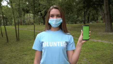 female volunteer in covid face mask holding smartphone with green screen. portrait of young beautiful caucasian woman posing with chromakey phone outdoors in summer park