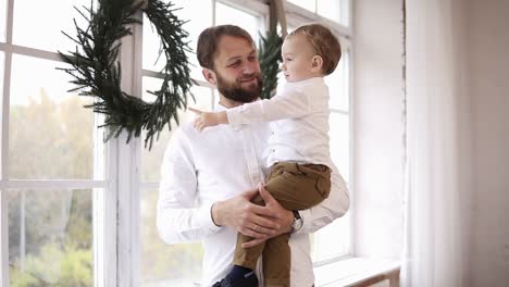 el joven padre está sosteniendo a su bebé, de pie junto a la ventana y mirando hacia afuera. la ventana está decorada con una corona de navidad