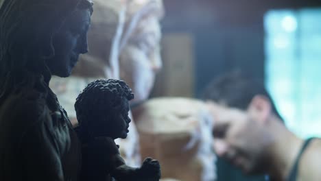 A sculptor works with focus as a clay model and a wooden statue of Madonna and Child stand side by side, symbolizing the transition from design to sacred creation