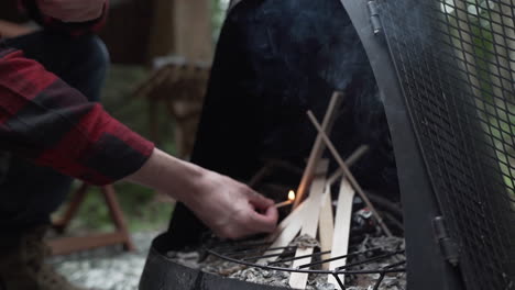 Ein-Junger-Erwachsener-Mann-Zündet-Ein-Streichholz-An-Und-Bringt-Es-Zu-Einem-Kleinen-Holzhaufen-In-Einer-Schwarzen-Feuerstelle,-Um-Neben-Einer-Hütte-Im-Wald-Ein-Feuer-Anzuzünden