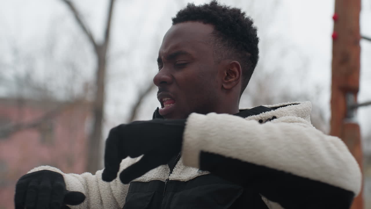 Young man performing fitness exercise outdoors in winter, standing in front of wooden workout equipment with snow-covered ground, focusing on his fitness routine despite cold weather
