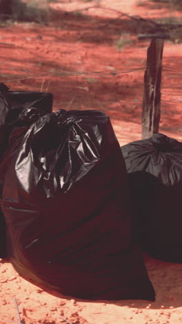 three black garbage bags sit beside a red dirt road and a wire fence.