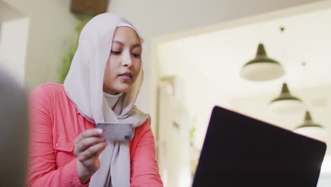 Video-Einer-Lächelnden-Gemischtrassigen-Frau-Im-Hijab,-Die-Zu-Hause-Mit-Laptop-Und-Kreditkarte-Online-Bezahlt