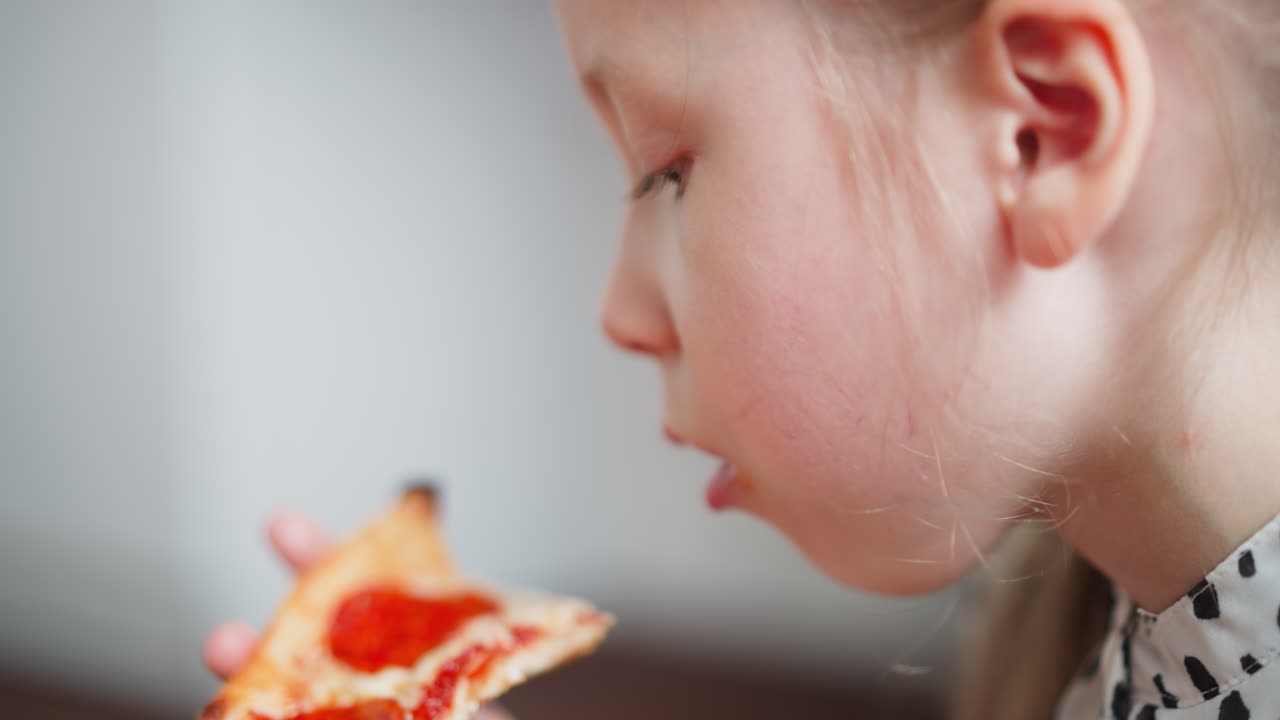 Close up of young girl wearing spotted clothes with hair tied back enjoying bite of pizza indoors, focusing on eating delicious snack with relaxed posture and soft natural light in cozy environment