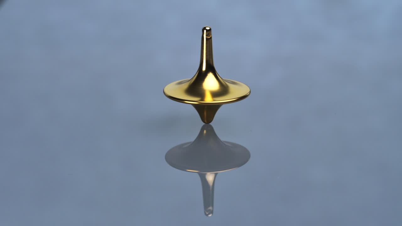 A polished golden top (totem) spins on a reflective grey-blue surface, creating a clear mirror image