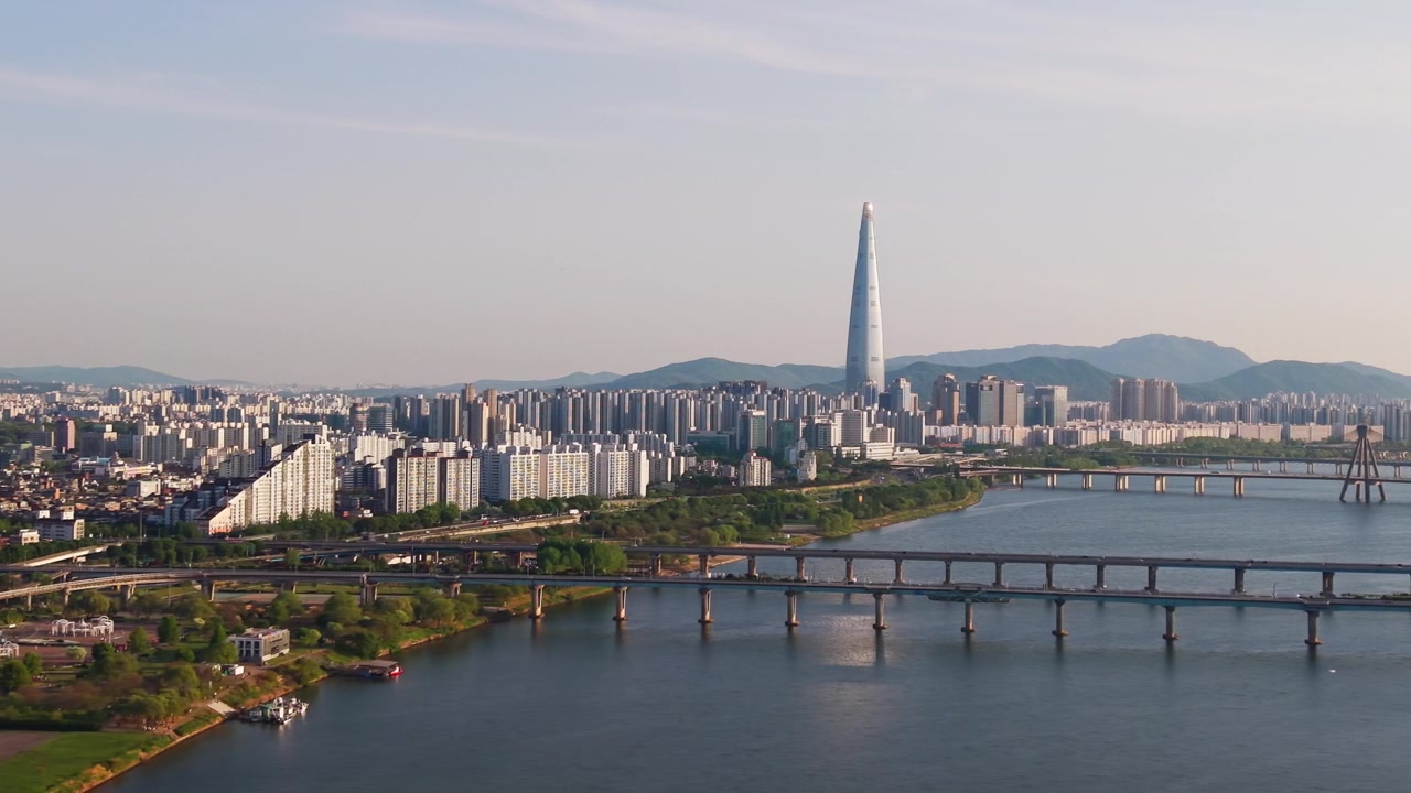 Un volo aereo laterale lungo il Parco Gwangnaru Hangang rivela i ponti Gwangjin e Cheonho sul fiume Han, con la Lotte World Tower nitidamente centrata sullo skyline primaverile limpido di Seul