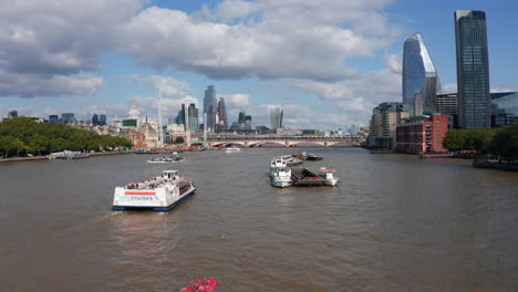 Fly-over-boats-cruising-on-River-Thames-in-Blackfriars.-Sunny-day-in-city.-Modern-luxurious-tall-office-buildings-in-background.-London,-UK