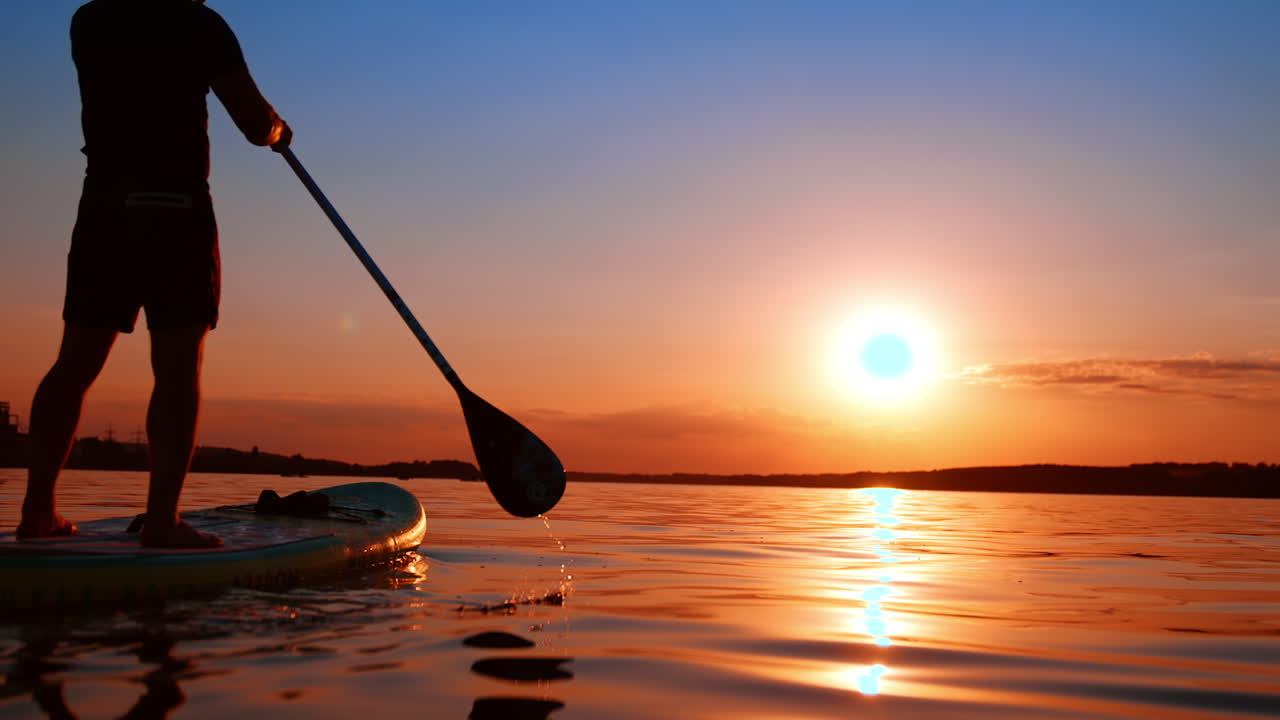 Man in cap rowing with an oar to move on his sup board by the river. Doing sports at sunset in beautiful nature. Low angle view.