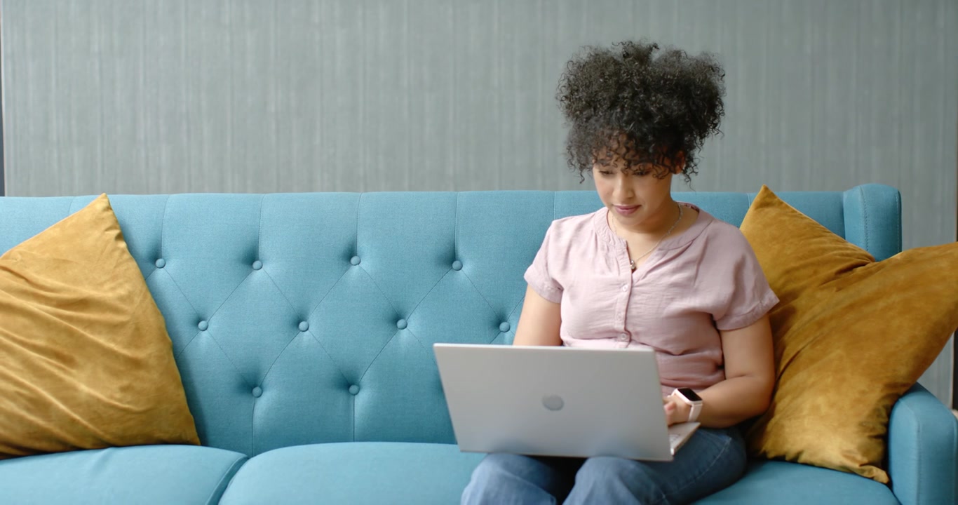 With laptop open, African American woman typing on laptop on teal tufted sofa, studying copy space