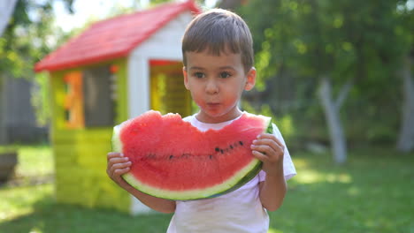 cute baby boy bites a big slice of ripe fresh watermelon and juice runs by the kid's chin. toddler stands up and shoves the piece close to camera.