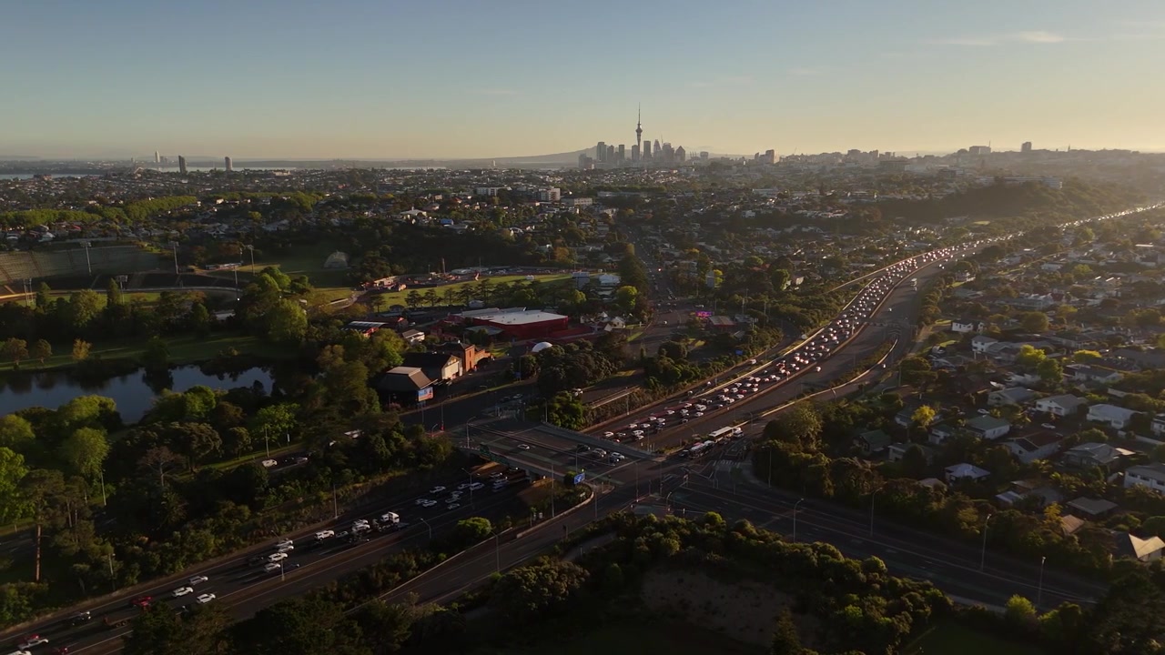 Veduta aerea a volo d'uccello di Auckland all'alba, con la Sky Tower, sobborghi estesi, traffico autostradale e una calda luce dorata mattutina.