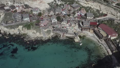 a drone orbits high above popeye village, overlooking the colorful film-set houses, winding road, turquoise anchor bay, and dramatic maltese coastline from an elevated perspective