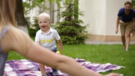 lächelnder vater mit seinem niedlichen, kleinen sohn. der junge rennt zu seiner mutter. fröhlich. die junge mutter umarmt den blonden jungen, küsst und umarmt ihn. dann geht er in die arme seines vaters. picknickkonzept.