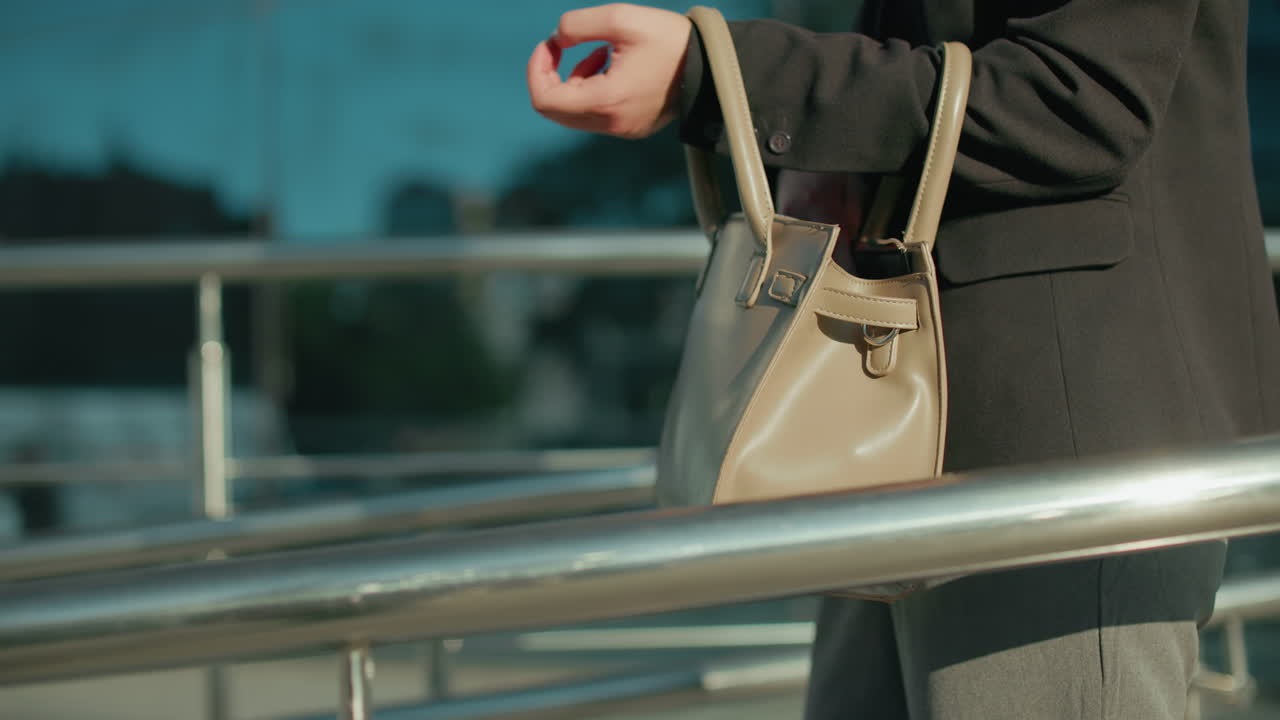 Close up side view of white lady preparing for lecture, checking handbag to retrieve keys, near modern glass building reflecting her, metal railings surrounding walkway