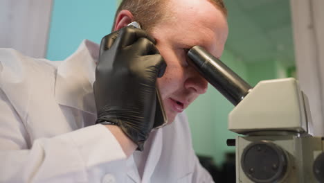 una vista de cerca de un hombre con una bata blanca de laboratorio y guantes negros, hablando por teléfono mientras observa de cerca a través de un microscopio en un entorno de laboratorio