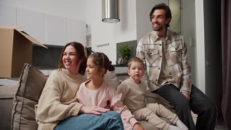 Glückliche-Familie-Auf-Dem-Sofa-Nach-Dem-Umzug-In-Eine-Neue-Moderne-Wohnung-Kommuniziert.-Ein-Brünetter-Mann-Mit-Stoppeln-In-Einem-Karierten-Beigen-Hemd,-Seine-Brünette-Frau,-Ihre-Kleine-Tochter-In-Rosa-Kleidung-Und-Ihr-Sohn-Sitzen-Auf-Dem-Sofa,-Nachdem-Sie-In-Eine-Neue-Moderne-Wohnung-Gezogen-Sind