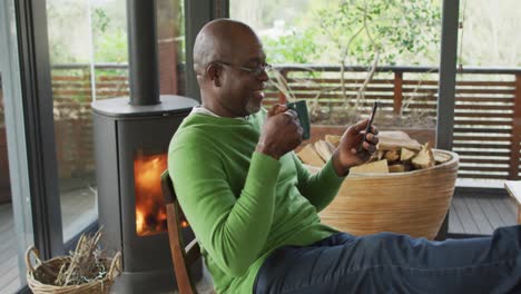 smiling african american senior man relaxing with feet up, using smartphone and drinking coffee