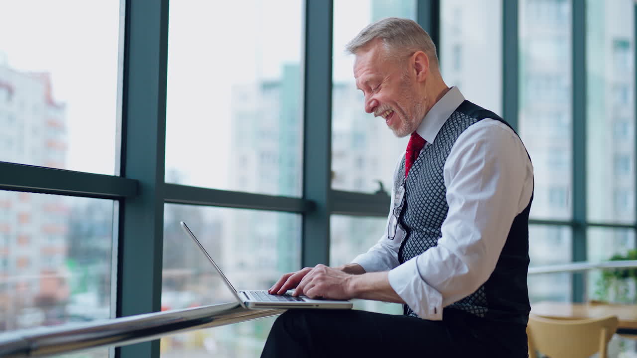Happy senior man typing on laptop near the big window. Excited freelancer working on a computer at home. Remote freelance work on retirement. Active modern lifestyle of older people.