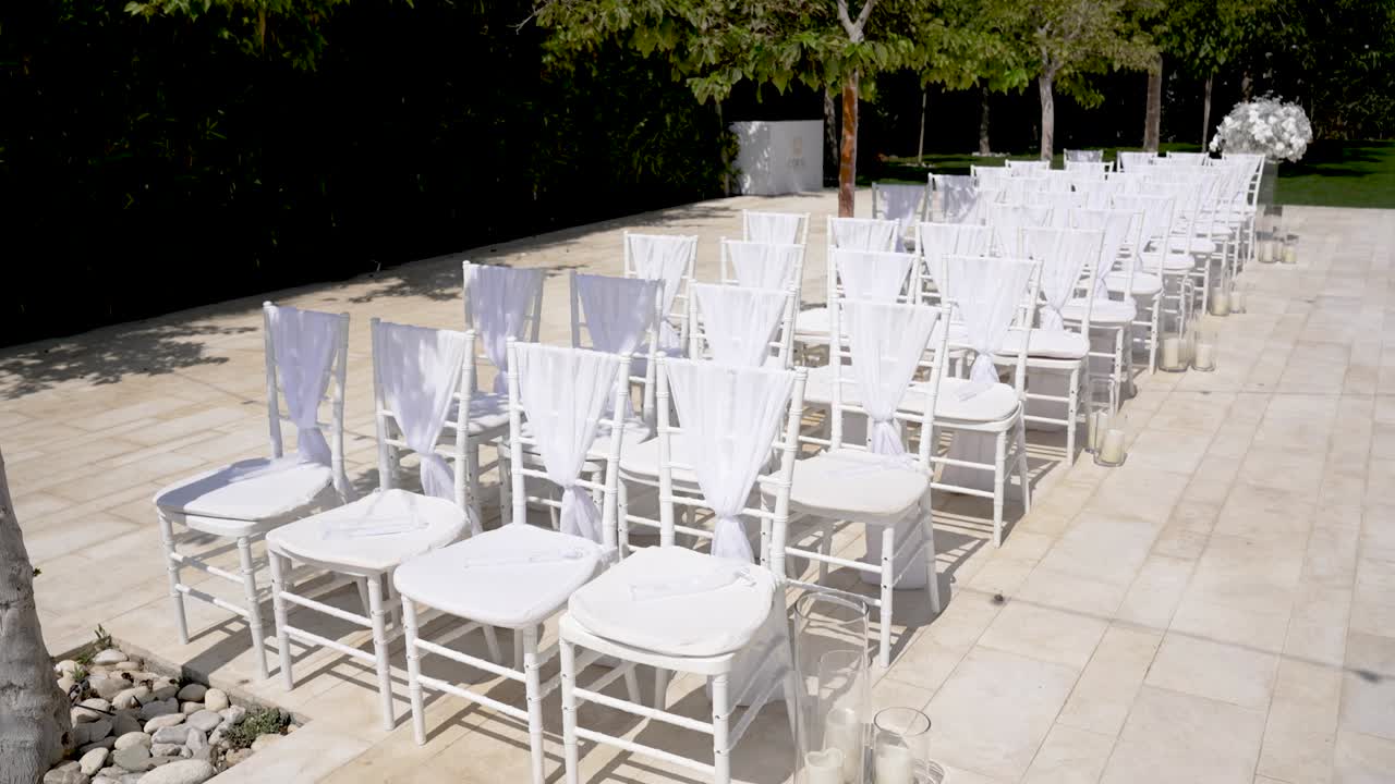 Rows of white chairs are arranged neatly in a beautiful garden for a wedding ceremony. The setting features peaceful greenery and simple floral accents, creating a romantic atmosphere